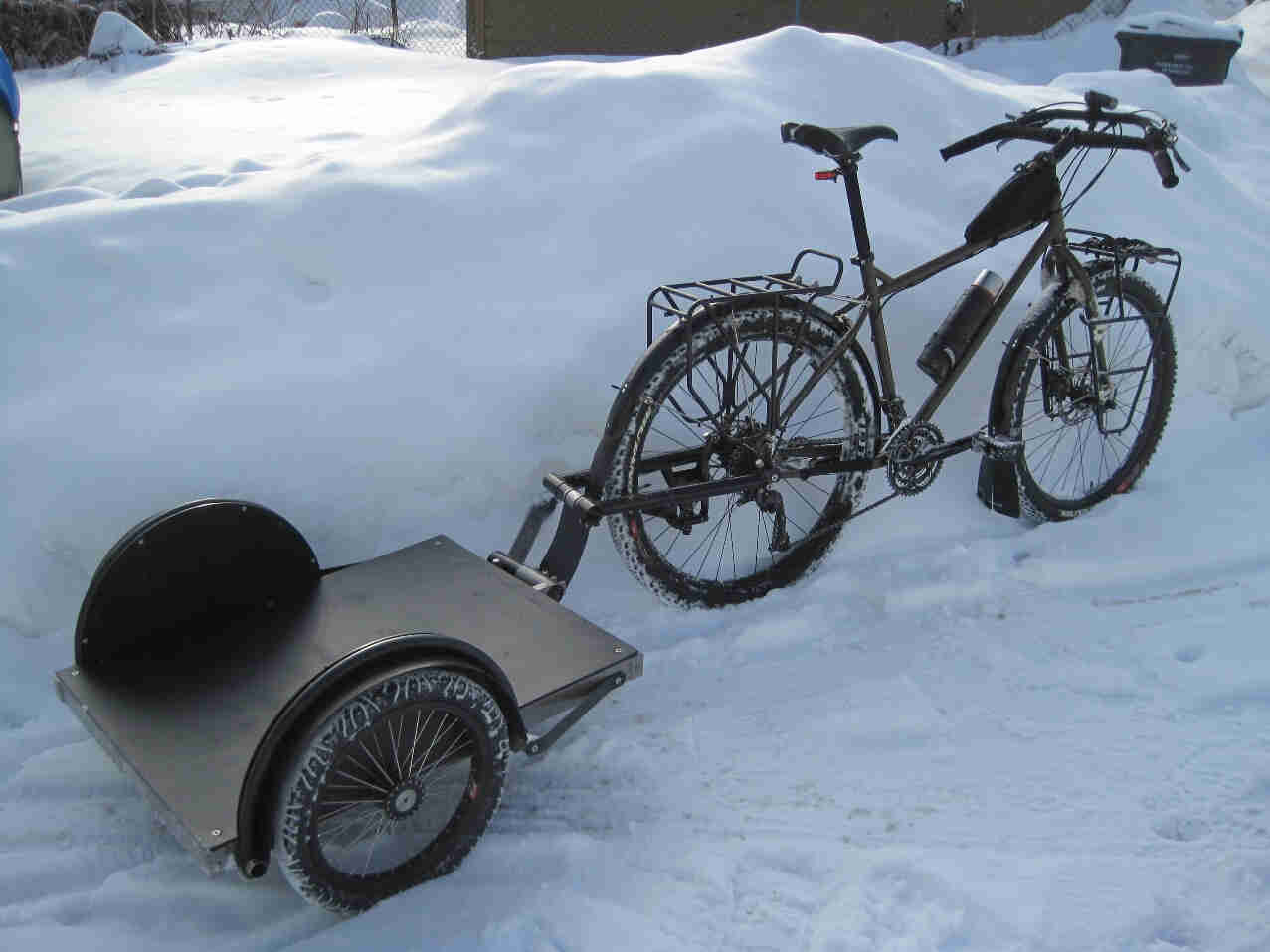 Right side view of a Surly Troll bike with a bike trailer hitched back on back, parked against a high snow bank